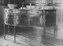 This fine Sheraton sideboard shows curved doors, and knife boxes with oval inlay of satinwood. The center cupboard is straight. The legs are reeded.