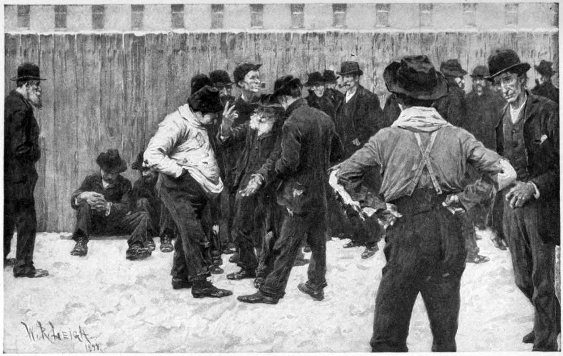 Men gathered outside. A fence is visible behind them with a wide building visible behind the fence.